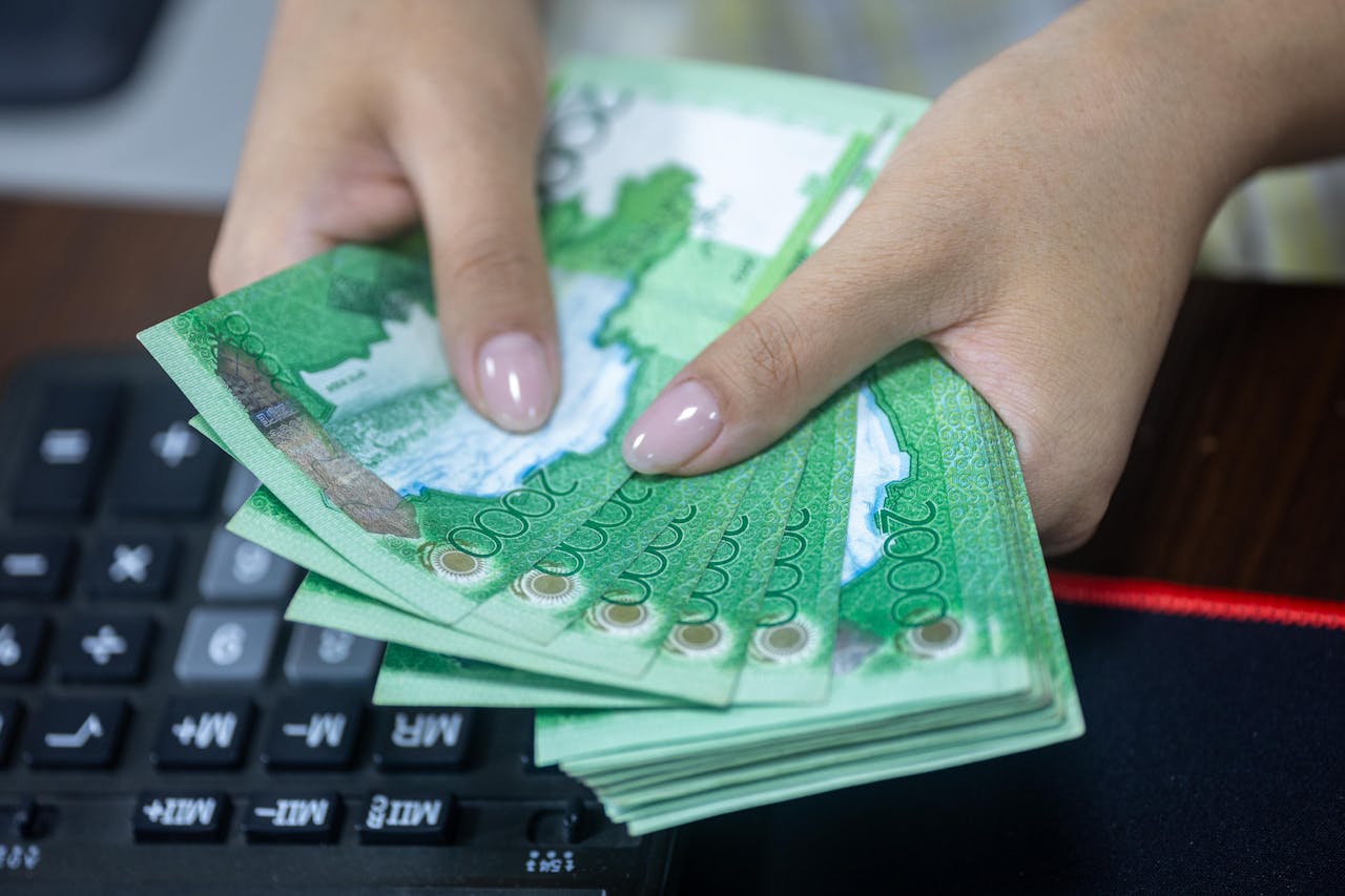 Close-up of hands counting Kazakhstani tenge bills beside a keyboard.